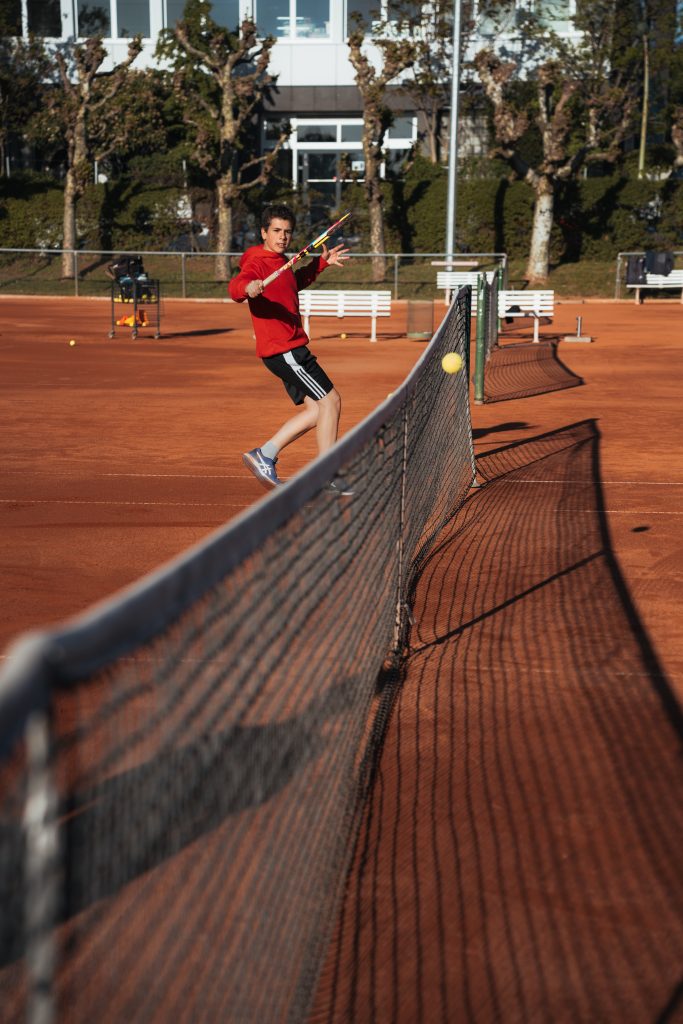 Jugendlicher im roten Shirt schlägt beim Tennis auf einem Sandplatz eine Vorhand am Netz