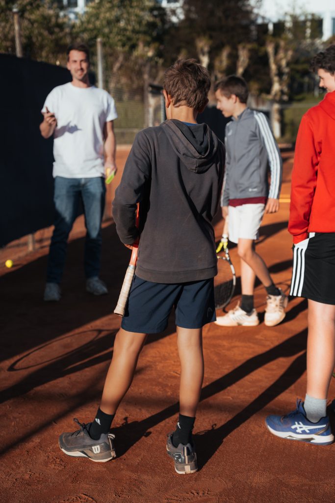 Tennistrainer im weißen Shirt spricht auf einem Sandplatz mit einer Gruppe Jugendlicher mit Schlägern