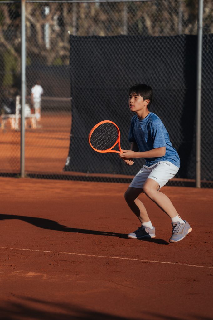 Junge im blauen Shirt steht mit orangefarbenem Tennisschläger in Bereitschaftshaltung auf einem Sandplatz